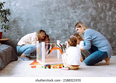 Attentive Little Girls Sitting On Haunches On Art Lesson, Painting With Women Mothers Teachers With Brushes On Stretch Film Wrapping Around Turned Table Legs In Grey Room. Early Development