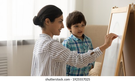 Attentive little indian boy preschooler take painting class from young millennial woman art teacher. Interested small child son watch inspired mother drawing picture on white board with colored chalks