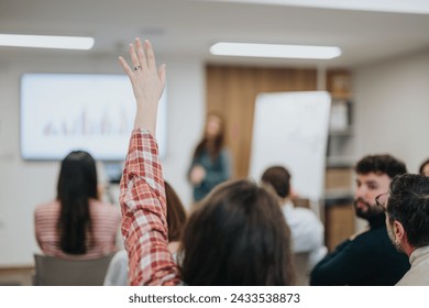 An attentive participant raises their hand to ask a question during a business seminar, with fellow attendees and presenter in the background. - Powered by Shutterstock