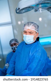 Attentive Professional Doctor Checks Operation Process In Modern Operating Room. Surgeon In Blue Scrubs, Hat And Facial Mask In Clinic. Portrait.