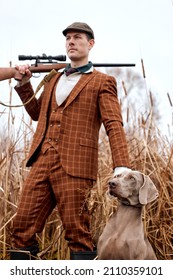 Attentive Strong Nice Hunting Dog Weimaraner Waiting To Get Command From Handsome Caucasian Gamekeeper Owner Man In Trendy Stylish Brown Suit, Walking In Rural Area Place. Outdoor Portrait.