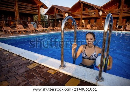 Attentive tanned pensive girl in a leopard swimsuit with a bright multi-colored inflatable ball in her hands climbs the stairs, getting out of a deep blue pool with clear transparent water