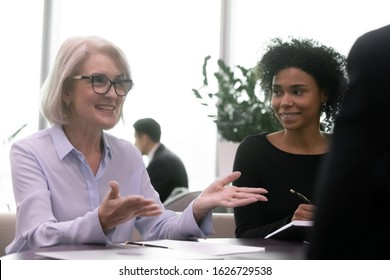 Attentive Young African American Assistant Listening To Smiling Middle Aged Female Boss In Eyeglasses Conducting Meeting With Partners, Discussing Contract Terms, Explain Agreement Details To Clients.