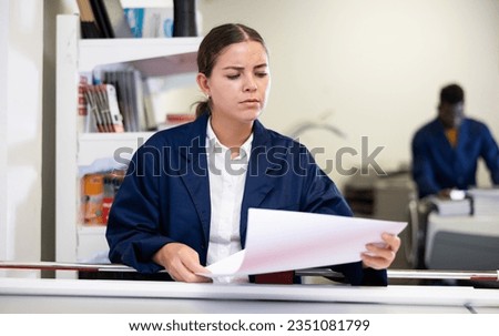 Attentive young female typographer in a blue uniform verifying printed sheet during work in the printer shop