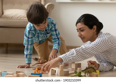 Attentive young indian nanny involve little boy in interesting game with toy dinosaurs and colorful blocks. Single hindu mother play with child son on warm floor teach to create story of fantasy world