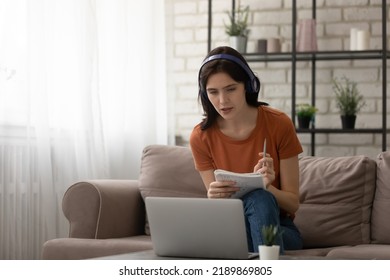 Attentive young woman sitting on couch by laptop listening to learning material in headphones. Female student study from home online at quarantine period taking notes of lecture to paper exercise - Shutterstock ID 2189869805