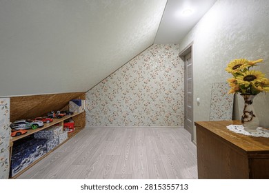 attic space has a slanted ceiling, floral wallpaper, light wood flooring, a wooden cabinet with sunflowers in a vase, a built‑in shelf with toys, and a floral‑patterned sofa