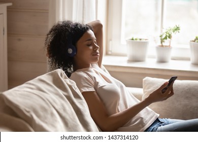 Attractive 30s mixed race woman wearing headphones closed her eyes holding smart phone listen enjoy favourite music lying in living room resting on couch spend lazy weekend at home, mood hobby concept