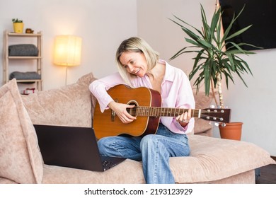 Attractive 40s Woman Plays The Guitar At Home. Smiling Adult Girl Takes Online Lesson On A Laptop Computer