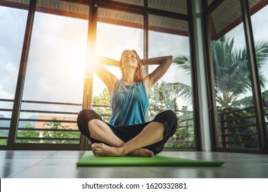 An attractive 40-year-old middle-aged woman practices yoga in a panoramic window room overlooking the garden at dawn in the sunshine. Meditation mindfulness healthy lifestyle concept. - Powered by Shutterstock