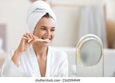 Attractive adult lady brushing teeth after shower, looking at mirror in bathroom, wearing white silky bathrobe and turban on her head, panorama with copy space. Oral care, dental health concept - Powered by Shutterstock