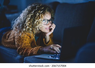 Attractive Adult Woman Smile And Use Laptop Computer And Internet Connection By Night Laying On The Sofa At Home. People Addicted To Technology And Social Media. Happy Female Lifestyle