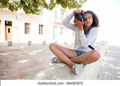 Attractive African American Black Teenager Girl Sitting On The Bench Of A Village Street, Using A Slr Digital Camera Taking Photographs On A Summer Holiday. Photography Student And Tourism Lifestyle.