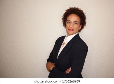 Attractive African American business woman with natural hair wearing a dark suit and white shirt standing confidently with arms crossed and smiling. Neutral background.