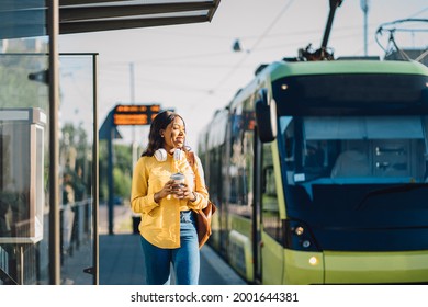 Attractive african american female traveler or student waiting for public transport standing on ram or troain stop outdoor at sunny morning with transport on blurred background.