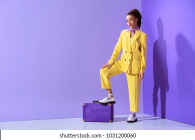 attractive african american girl posing in yellow suit with purple suitcase, on trendy ultra violet background