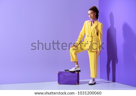 attractive african american girl posing in yellow suit with purple suitcase, on trendy ultra violet background