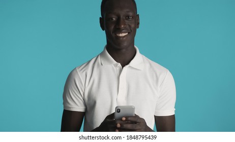 Attractive African American Guy Dressed In Casual Wear Getting Message On Smartphone Friendly Smiling In Camera Isolated On Blue Background