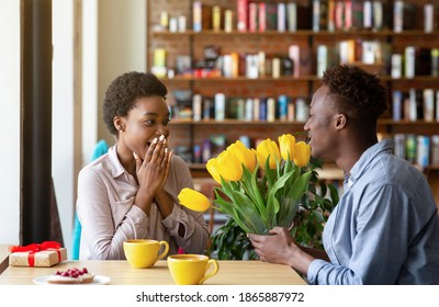 Attractive African American guy presenting his girlfriend with beautiful tulips, expressing his love at city cafe. Romantic black couple celebrating anniversary with gifts and flowers at coffee shop