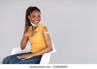 Attractive African American Lady Smiling And Pointing At Band Aid After Receiving Coronavirus Vaccine Shot On Grey Background, Copy Space. Millennial Black Woman Getting Vaccinated Against Covid