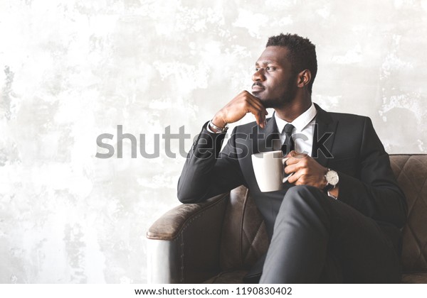 スタイリッシュなインテリアに ビジネススーツを着た魅力的なアフリカ系アメリカ人男性 ビジネスマンと企業リーダー 人は仕事の前に朝のコーヒーを飲んで元気を出す の写真素材 今すぐ編集