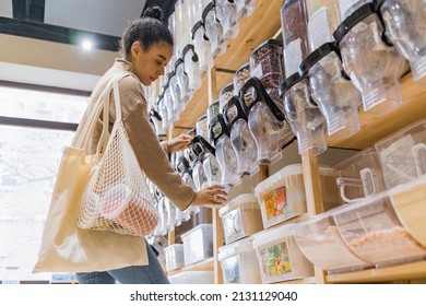 Attractive african american woman buying organic cereals in sustainable zero waste grocery store. Young woman with cotton bags refilling reusable container with cereals in local grocery store
