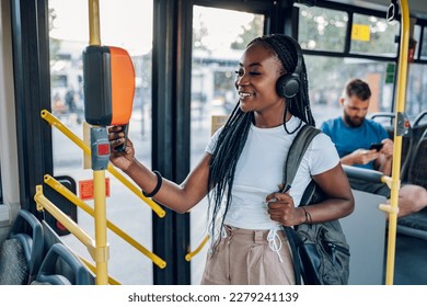 Attractive african american woman paying a bus ticket with her smartphone. Young beautiful woman using public transportation and standing in the bus during a ride. Contactless payment.