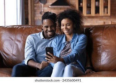 Attractive African couple sit on sofa looking at cellphone screen, make video call use mobile app, watch internet content, taking selfie pictures, spend leisure at home enjoy modern tech usage concept - Powered by Shutterstock