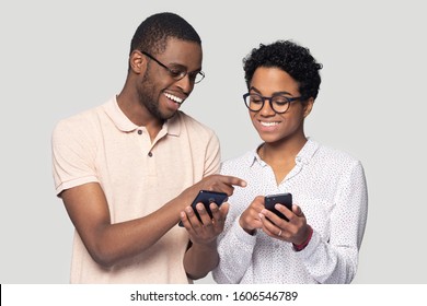 Attractive african ethnicity couple hold smart phones having fun in internet, guy help explain to girl new cool free online application, point finger show to photos, isolated on gray studio background