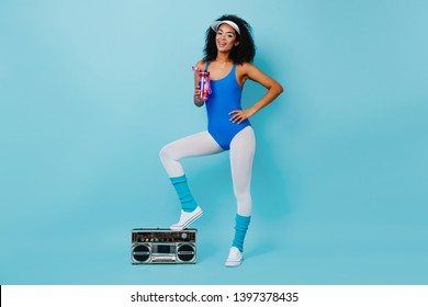 Attractive african lady in aerobics outfit standing on boombox. Studio shot of wonderful black girl drinking water after training.