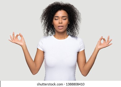 Attractive African Millennial Woman In White T-shirt Doing Yoga Exercise Girl Meditating Standing Over Grey Background Closed Eyes Feels Internal Balance Make Fingers Mudra Gesture, No Stress Concept