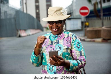 Attractive African Woman Standing Showing Thumb Up, Holding A Mobile Phone, Expressing Her Sincere Emotions And Wearing A Hat While Smiling With Gestures.