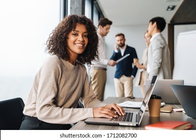 Attraktive afrikanische junge selbstbewusste Geschäftsfrau sitzt am Bürotisch mit einer Gruppe von Kollegen im Hintergrund, arbeitet am Laptop-Computer