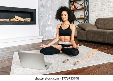 Attractive African-American woman is practicing yoga with video tutorials, she sits on the floor in lotus pose with eyes closed in front of laptop - Powered by Shutterstock