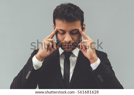 Attractive Afro American businessman in classic suit is touching his temples while concentrating, on gray background