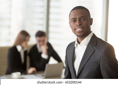 Attractive afro american smiling businessman looking at camera standing in foreground, young cheerful african leading manager posing with subordinates at the background, multi-ethnic team portrait