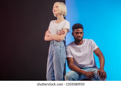 Attractive Afro Man Is Sitting On The Chair And Looking At The Camera, While His Caucasian Albino Girlfriend Standing With Crossed Arms Bear Him, Copy Space