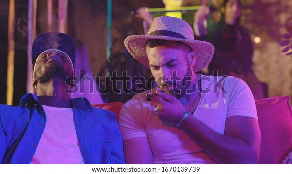 Attractive\
afro-american man smoking weed with a pipe enjoying high euphoria\
chilling with friend at the\
party.