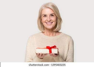 Attractive aged blond woman looks at camera holding presents gift box red bow package feels happy studio head shot isolated on grey background. Life events celebration congratulation love care concept - Powered by Shutterstock