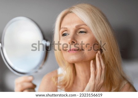 Attractive aged european woman looking in magnifying mirror and touching her face smooth skin, beautiful senior lady enjoying her appearance, closeup