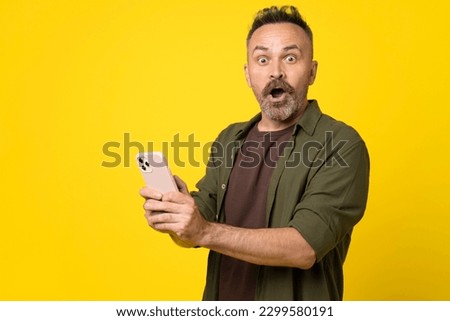 attractive amazed bristle man using digital smartphone in hands demonstrating shock from news or sms, reaction influencer post review isolated over yellow background