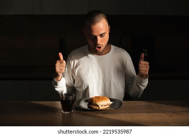 Attractive Amazed Surprised Man Show Thumbs Up Gesture And Sitting Near Table With Plate With Burger. Fast Food. Male Wear White Long Sleeves Sitting At House In Kitchen And Look At Burger, Open Mouth