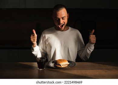 Attractive Amazed Surprised Man Show Thumbs Up Gesture And Sitting Near Table With Plate With Burger. Fast Food. Male Wear White Long Sleeves Sitting At House In Kitchen And Look At Burger, Open Mouth