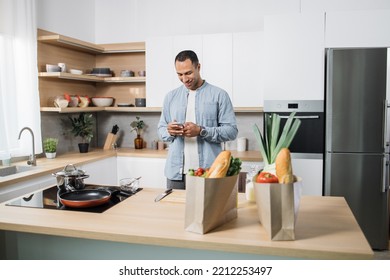 Attractive Arab Man Preparing Romantic Dinner On Home Kitchen. Serious Concentrated Guy Looking For Good Recipe At Smart Phone. Healthy Food Menu App. Cookbook Mobile Application. Technology And Home