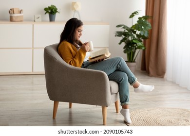 Attractive arab woman with cup of aromatic coffee and open book relaxing in armchair at home, having lazy weekend indoors. Young lady enjoying hot beverage and reading exciting story