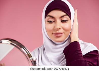 Attractive Arab Woman In Hijab Looks In Mirror. Isolated On Pink Background. Studio Portrait.