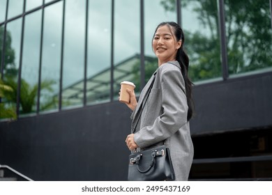 An attractive Asian businesswoman in a fashionable suit is smiling at the camera, holding a takeaway coffee cup, walking in the city, going to work in the morning. - Powered by Shutterstock