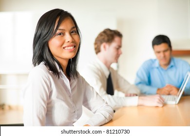 Attractive Asian Businesswoman Looking At The Camera Sitting At Conference Room Table In Meeting With Diverse Group Of Business People Including A Latino And Caucasian Male. Horizontal