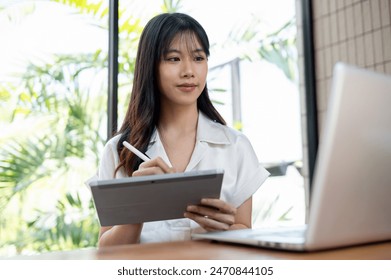 An attractive Asian businesswoman multitasks, using her digital tablet and laptop computer while working on her project in a modern office or co-working space. - Powered by Shutterstock