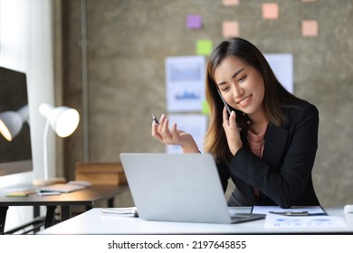 Attractive Asian Businesswoman Talking On Mobile Phone Working  In Modern Office. Happy Business Woman Talking On Mobile Phone While Analyzing Monthly Work Schedule In Laptop.